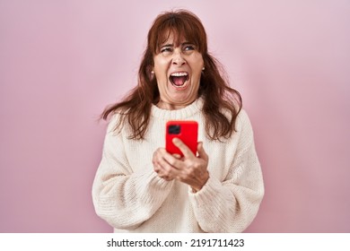 Middle Age Hispanic Woman Using Smartphone Typing Message Angry And Mad Screaming Frustrated And Furious, Shouting With Anger Looking Up. 