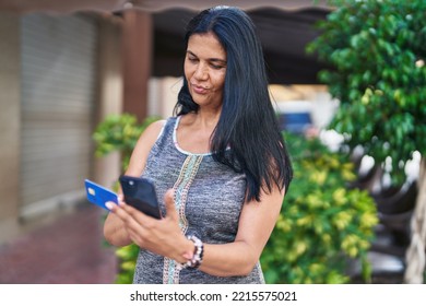 Middle Age Hispanic Woman Using Smartphone And Credit Card With Serious Expression At Street