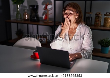 Middle age hispanic woman using laptop at home at night bored yawning tired covering mouth with hand. restless and sleepiness. 