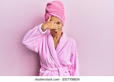 Middle Age Hispanic Woman Wearing Shower Towel Cap And Bathrobe Peeking In Shock Covering Face And Eyes With Hand, Looking Through Fingers With Embarrassed Expression. 