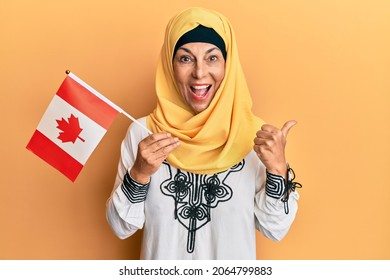 Middle Age Hispanic Woman Wearing Hijab Holding Canada Flag Pointing Thumb Up To The Side Smiling Happy With Open Mouth 