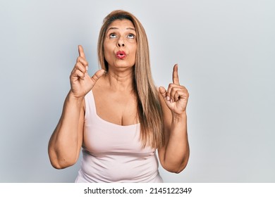 Middle Age Hispanic Woman Wearing Casual Style With Sleeveless Shirt Amazed And Surprised Looking Up And Pointing With Fingers And Raised Arms. 