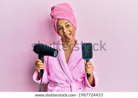 Middle age hispanic woman wearing bathrobe using hair comb and dryer sticking tongue out happy with funny expression. 