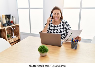 Middle Age Hispanic Woman Working At The Office With Laptop Speaking On The Phone Sticking Tongue Out Happy With Funny Expression. 