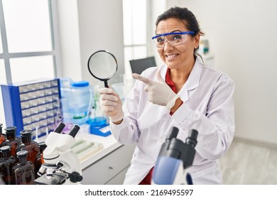 Middle Age Hispanic Woman Working At Scientist Laboratory Holding Magnifying Glass Smiling Happy Pointing With Hand And Finger 