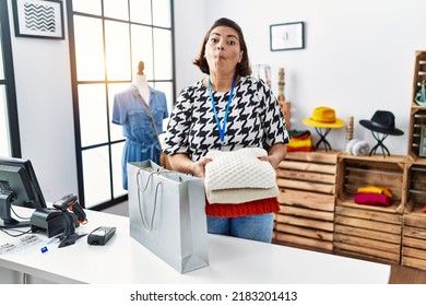 Middle Age Hispanic Woman Working At Retail Boutique Putting Clothes Inside Bag Making Fish Face With Mouth And Squinting Eyes, Crazy And Comical. 