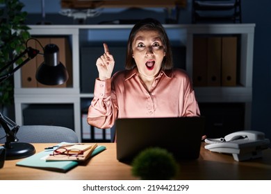 Middle Age Hispanic Woman Working Using Computer Laptop Late At Night Pointing Finger Up With Successful Idea. Exited And Happy. Number One. 