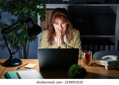 Middle Age Hispanic Woman Working Using Computer Laptop At Night Praying With Hands Together Asking For Forgiveness Smiling Confident. 