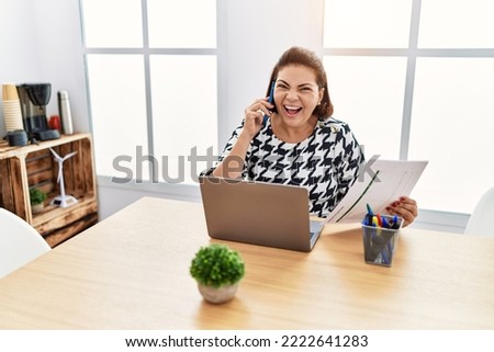 Middle age hispanic woman working at the office with laptop speaking on the phone smiling and laughing hard out loud because funny crazy joke. 