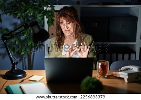 Middle age hispanic woman working using computer laptop at night disgusted expression, displeased and fearful doing disgust face because aversion reaction. with hands raised 