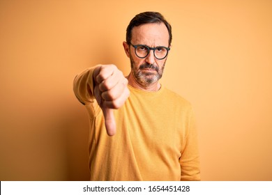 Middle age hoary man wearing casual sweater and glasses over isolated yellow background looking unhappy and angry showing rejection and negative with thumbs down gesture. Bad expression.