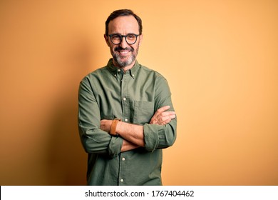 Middle age hoary man wearing casual green shirt and glasses over isolated yellow background happy face smiling with crossed arms looking at the camera. Positive person.