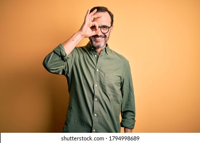 Middle Age Hoary Man Wearing Casual Green Shirt And Glasses Over Isolated Yellow Background Doing Ok Gesture With Hand Smiling, Eye Looking Through Fingers With Happy Face.