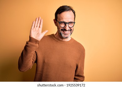 Middle age hoary man wearing brown sweater and glasses over isolated yellow background Waiving saying hello happy and smiling, friendly welcome gesture