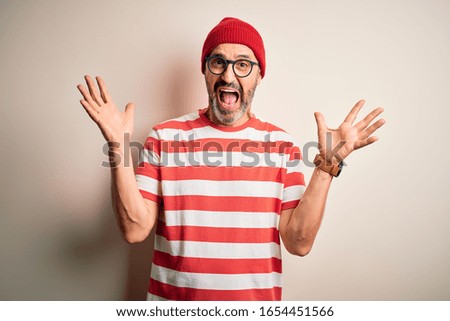 Middle age hoary man wearing striped t-shirt glasses and cap over isolated white background celebrating crazy and amazed for success with arms raised and open eyes screaming excited. Winner concept