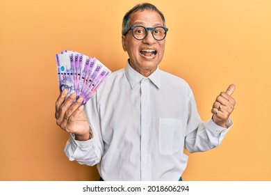 Middle Age Indian Man Holding 100 Philippine Peso Banknotes Pointing Thumb Up To The Side Smiling Happy With Open Mouth 