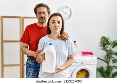 Middle Age Interracial Couple Doing Laundry Holding Detergent Bottle Thinking Attitude And Sober Expression Looking Self Confident 