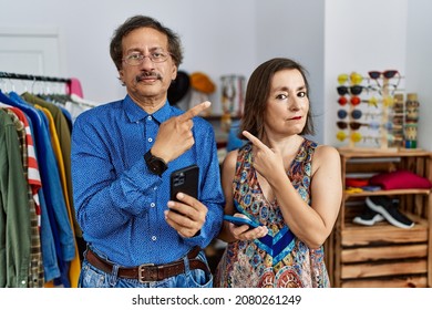 Middle Age Interracial Couple At Retail Shop Using Smartphone Pointing With Hand Finger To The Side Showing Advertisement, Serious And Calm Face 