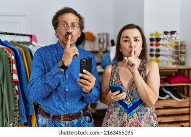 Middle Age Interracial Couple At Retail Shop Using Smartphone Asking To Be Quiet With Finger On Lips. Silence And Secret Concept. 