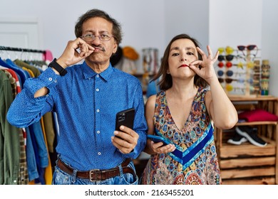 Middle Age Interracial Couple At Retail Shop Using Smartphone Mouth And Lips Shut As Zip With Fingers. Secret And Silent, Taboo Talking 