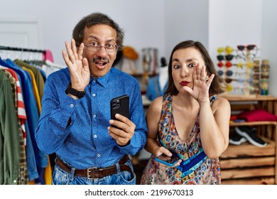 Middle Age Interracial Couple At Retail Shop Using Smartphone Hand On Mouth Telling Secret Rumor, Whispering Malicious Talk Conversation 