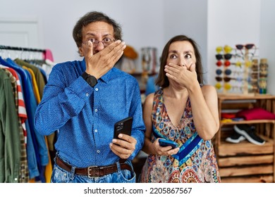 Middle Age Interracial Couple At Retail Shop Using Smartphone Shocked Covering Mouth With Hands For Mistake. Secret Concept. 