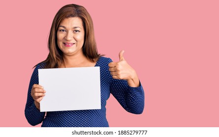 Middle age latin woman holding paper banner with blank space smiling happy and positive, thumb up doing excellent and approval sign 