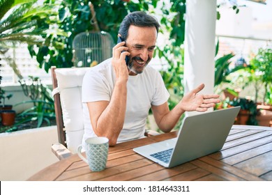 Middle Age Man With Beard Smiling Happy At The Terrace Working From Home Using Laptop Speaking On The Phone