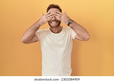 Middle age man with beard standing over yellow background covering eyes with hands smiling cheerful and funny. blind concept.  - Powered by Shutterstock