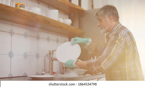 Middle Age Man With Beard Wearing Gloves Washing Dishes At Home. Side View Of Senior Male Cleaning Plates After Dinner In Washbasin. Mature Man Doing Household