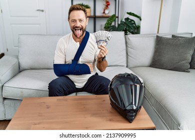 Middle age man with beard wearing arm on sling for motorbike accident holding insurance money sticking tongue out happy with funny expression. 