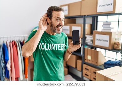 Middle Age Man With Beard Wearing Volunteer T Shirt Holding Smartphone Smiling With Hand Over Ear Listening An Hearing To Rumor Or Gossip. Deafness Concept. 