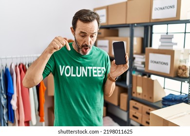 Middle Age Man With Beard Wearing Volunteer T Shirt Holding Smartphone Pointing Down With Fingers Showing Advertisement, Surprised Face And Open Mouth 