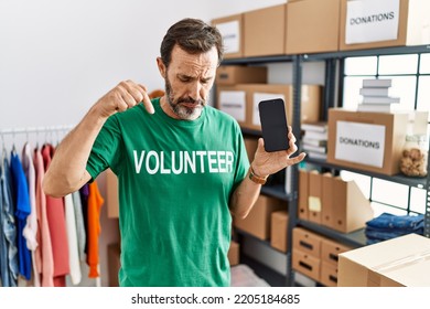 Middle Age Man With Beard Wearing Volunteer T Shirt Holding Smartphone Pointing Down Looking Sad And Upset, Indicating Direction With Fingers, Unhappy And Depressed. 