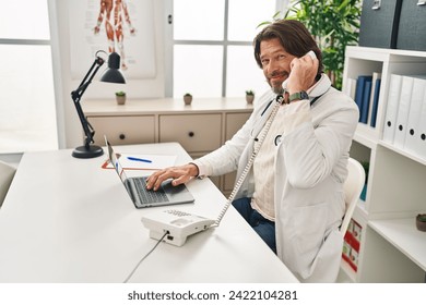 Middle age man doctor using laptop talking on telephone at clinic - Powered by Shutterstock