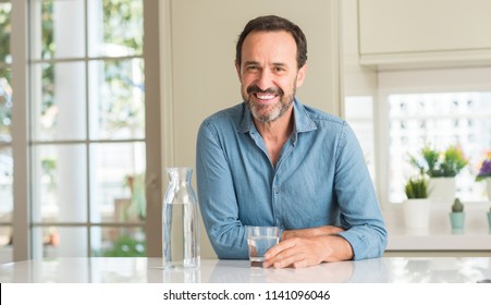 Middle age man drinking a glass of water with a happy face standing and smiling with a confident smile showing teeth