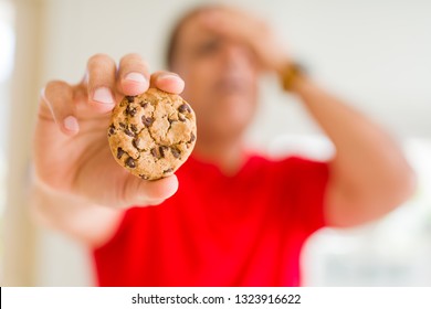 Middle age man eating chocolate chips cookies at home stressed with hand on head, shocked with shame and surprise face, angry and frustrated. Fear and upset for mistake.