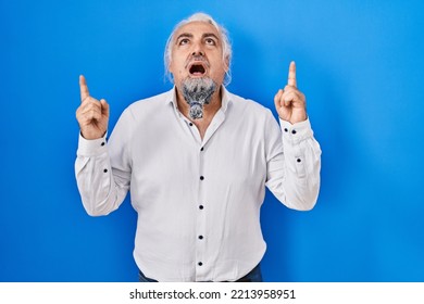 Middle Age Man With Grey Hair Standing Over Blue Background Amazed And Surprised Looking Up And Pointing With Fingers And Raised Arms. 