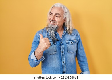 Middle Age Man With Grey Hair Standing Over Yellow Background Smiling With Happy Face Looking And Pointing To The Side With Thumb Up. 