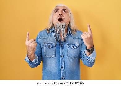 Middle Age Man With Grey Hair Standing Over Yellow Background Amazed And Surprised Looking Up And Pointing With Fingers And Raised Arms. 