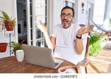 Middle Age Man Using Computer Laptop At Home Clueless And Confused Expression With Arms And Hands Raised. Doubt Concept. 