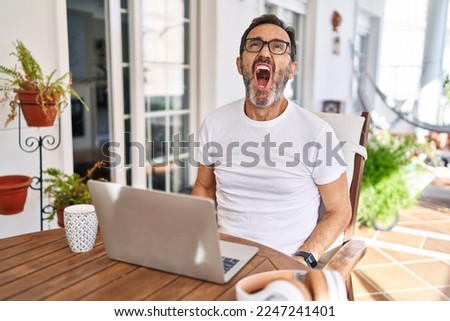 Middle age man using computer laptop at home angry and mad screaming frustrated and furious, shouting with anger. rage and aggressive concept. 