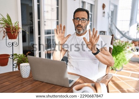 Middle age man using computer laptop at home moving away hands palms showing refusal and denial with afraid and disgusting expression. stop and forbidden. 