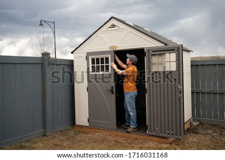 Middle age man wearing orange t shirt, jeans and baseball hat standing in doorway of his garden shed and fixing the door. A man finishes to build the backyard shed. Early spring garden work concept.
