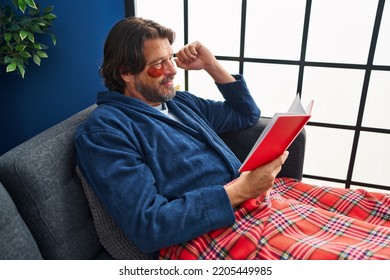 Middle Age Man Wearing Under Eye Patches Reading Book On Sofa At Home