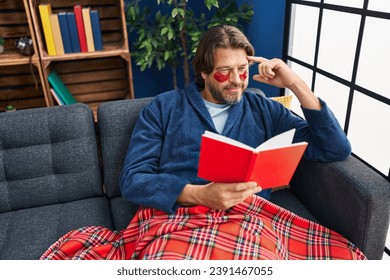 Middle age man wearing under eye patches reading book on sofa at home - Powered by Shutterstock