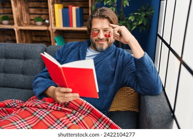 Middle age man wearing under eye patches reading book on sofa at home - Powered by Shutterstock