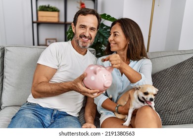 Middle Age Man And Woman Couple Holding Piggy Bank Sitting On Sofa With Dog At Home