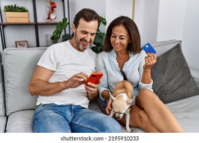 Middle Age Man And Woman Couple Using Smartphone And Credit Card Sitting On Sofa With Dog At Home