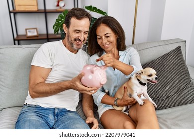 Middle Age Man And Woman Couple Holding Piggy Bank Sitting On Sofa With Dog At Home